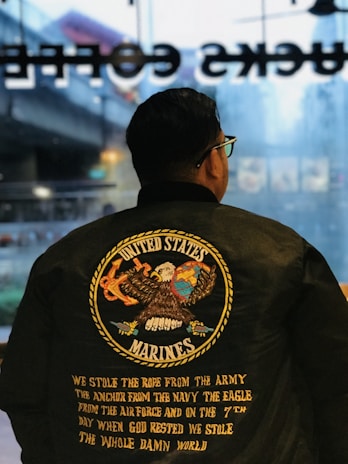 A person is wearing a black jacket with a large emblem on the back featuring an eagle holding a globe and an anchor. The text reads 'United States Marines' with additional text describing the military branches and a quote underneath.