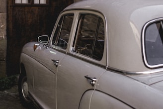 A vintage car with a cream-colored exterior is parked next to a wooden wall. The car's rounded body design and chrome accents reflect its classic style. The background features an aged, rustic setting with dim lighting, emphasizing the car's retro appearance.