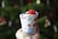 A hand holding a small, cylindrical gift wrapped in festive paper. The paper features red, white, and gray holiday-themed designs, including mittens and stars. A bright red ribbon is attached to the top of the gift. The background is blurred, with hints of green, likely indicating a Christmas tree.