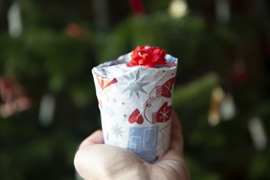 A hand holding a small, cylindrical gift wrapped in festive paper. The paper features red, white, and gray holiday-themed designs, including mittens and stars. A bright red ribbon is attached to the top of the gift. The background is blurred, with hints of green, likely indicating a Christmas tree.