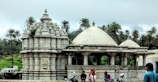 An ancient stone temple characterized by ornate carvings sits among lush greenery. Surrounding the structure are tall palm trees under an overcast sky. A group of people, including a woman in traditional attire, gather near the temple, some on scooters.