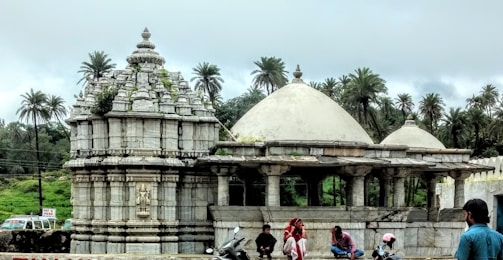 An ancient stone temple characterized by ornate carvings sits among lush greenery. Surrounding the structure are tall palm trees under an overcast sky. A group of people, including a woman in traditional attire, gather near the temple, some on scooters.