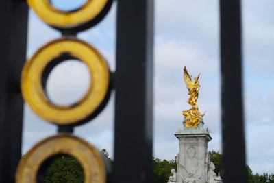 A golden statue of Victory is prominently displayed atop a tall, intricately decorated pedestal. The statue is viewed through an ornate fence with concentric circles, providing a sense of depth. The background features a cloudy sky and lush greenery.