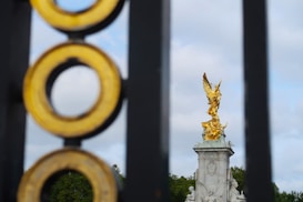 A golden statue of Victory is prominently displayed atop a tall, intricately decorated pedestal. The statue is viewed through an ornate fence with concentric circles, providing a sense of depth. The background features a cloudy sky and lush greenery.
