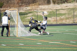 Three players wearing protective gear are engaged in a lacrosse game on a green, marked field. One player in a black and yellow uniform appears to be attempting to score, raising a lacrosse stick, while another player in white and black tries to block. A goalie stands in front of the goal, ready to defend.