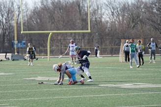 A lacrosse match is taking place on an outdoor field. Two players in action are at the center, one bending down and the other closely contesting the ball. In the background, other players and a goalpost are visible. Trees with bare branches and a clear sky surround the field.