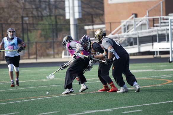 Several lacrosse players wearing helmets and padded gear are actively engaged in a game on a green turf field. Two players in the foreground are competing for control of the ball, which is on the ground. Other players are positioned in the background near the sidelines and goal area. A fence lines the edge of the field with some bleachers visible.