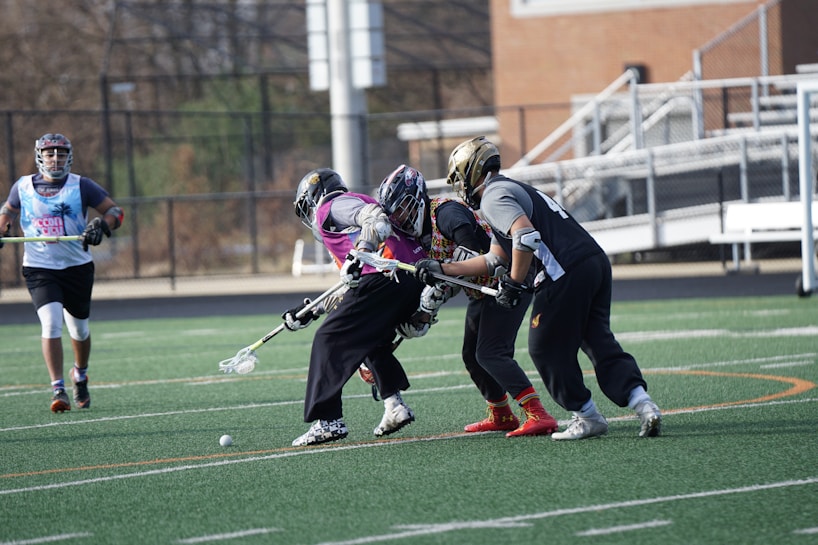 Several lacrosse players wearing helmets and padded gear are actively engaged in a game on a green turf field. Two players in the foreground are competing for control of the ball, which is on the ground. Other players are positioned in the background near the sidelines and goal area. A fence lines the edge of the field with some bleachers visible.