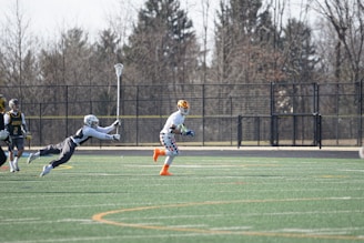 A group of athletes engaged in a sports game on a green field. One player is running forward with possession of the ball, wearing colorful socks and a checkered uniform, while another player in dark clothing dives to intercept or tackle. Other players are present in the background. Leafless trees and a metal fence surround the field.