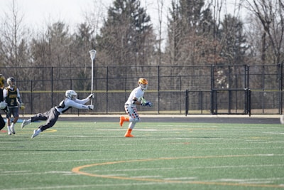 A group of athletes engaged in a sports game on a green field. One player is running forward with possession of the ball, wearing colorful socks and a checkered uniform, while another player in dark clothing dives to intercept or tackle. Other players are present in the background. Leafless trees and a metal fence surround the field.