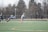 A group of athletes engaged in a sports game on a green field. One player is running forward with possession of the ball, wearing colorful socks and a checkered uniform, while another player in dark clothing dives to intercept or tackle. Other players are present in the background. Leafless trees and a metal fence surround the field.
