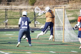 A group of lacrosse players actively engaged in a game. One player wearing a brown jersey is positioned close to the goal post, seemingly attempting to score while another player in a blue jersey marked with the number 14 approaches from behind. The scene takes place on a sports field marked with various lines.