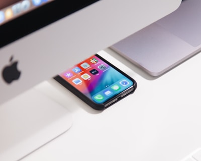 A smartphone with a colorful screen is positioned on a desk, partially under a computer. The image shows an Apple iMac and a MacBook, both with a sleek design, situated in an organized workspace.