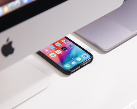 A smartphone with a colorful screen is positioned on a desk, partially under a computer. The image shows an Apple iMac and a MacBook, both with a sleek design, situated in an organized workspace.