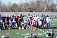 A group of people, primarily young men, posing together on a sports field. They appear to be a lacrosse team, with several individuals wearing sports attire and protective gear. Bleachers and trees can be seen in the background on a clear day.