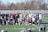 A group of people, primarily young men, posing together on a sports field. They appear to be a lacrosse team, with several individuals wearing sports attire and protective gear. Bleachers and trees can be seen in the background on a clear day.