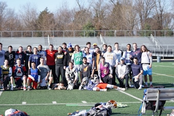 A group of people, primarily young men, posing together on a sports field. They appear to be a lacrosse team, with several individuals wearing sports attire and protective gear. Bleachers and trees can be seen in the background on a clear day.