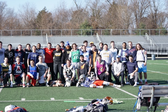 A group of people, primarily young men, posing together on a sports field. They appear to be a lacrosse team, with several individuals wearing sports attire and protective gear. Bleachers and trees can be seen in the background on a clear day.