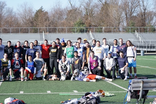 A group of people, primarily young men, posing together on a sports field. They appear to be a lacrosse team, with several individuals wearing sports attire and protective gear. Bleachers and trees can be seen in the background on a clear day.