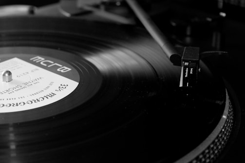 A black and white image of a vinyl record spinning on a turntable. The tonearm is positioned over the record, with the needle touching the grooves. The label on the vinyl displays text and details about the music. The scene is close-up and focuses on the physical texture of the record and the machinery of the turntable.