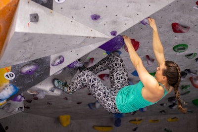 A woman is engaged in indoor rock climbing, gripping holds on a climbing wall with vibrant colors. She wears a turquoise tank top and patterned climbing pants. The climbing wall is filled with various holds and routes marked by different colors and numbers.