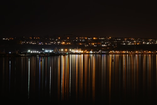 A nighttime urban landscape showcases a waterfront with numerous city lights reflecting off the calm water. The reflections create vertical streaks of various colors, producing a vibrant and dynamic visual effect.
