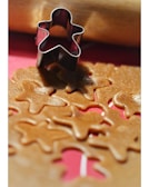 A metal gingerbread man-shaped cookie cutter is placed on a sheet of rolled-out dough that has several cutouts of the same shape. A wooden rolling pin can be seen in the background.