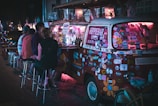 A vibrant, sticker-covered van is converted into a mobile bar with glowing pink lights. Several people are seated on stools beside the van enjoying drinks under the night sky. The van displays a sign indicating it is a 'Hippie Bar', open during evening hours. The atmosphere appears lively and informal.