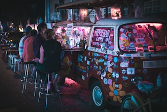 A vibrant, sticker-covered van is converted into a mobile bar with glowing pink lights. Several people are seated on stools beside the van enjoying drinks under the night sky. The van displays a sign indicating it is a 'Hippie Bar', open during evening hours. The atmosphere appears lively and informal.