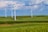 Several large wind turbines are situated across a sprawling green landscape, under a partly cloudy sky. The terrain is predominantly grassy with some hay bales scattered around, indicating an agricultural area.