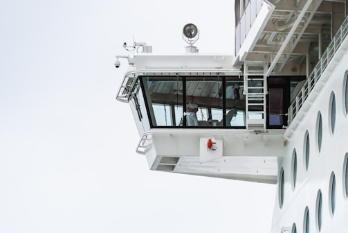 The image depicts the bridge of a large ship, prominently featuring the glass-enclosed command center where a person in a white uniform is visible. The exterior of the ship is a sleek white with modern design elements, including circular windows and safety equipment. The sky is overcast, giving the scene a clean and minimalistic look.