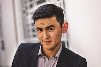 A young man with neatly styled dark hair is wearing a pinstriped suit and a light-colored shirt. He has a slight smile and is looking directly at the camera. The background is blurred, showing architectural elements with a light color scheme.