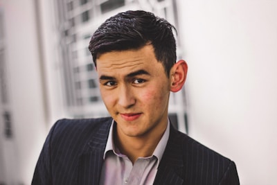 A young man with neatly styled dark hair is wearing a pinstriped suit and a light-colored shirt. He has a slight smile and is looking directly at the camera. The background is blurred, showing architectural elements with a light color scheme.