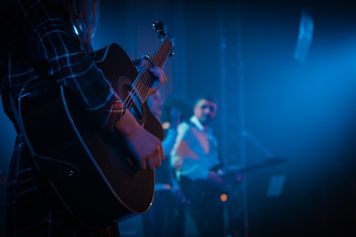A person playing an acoustic guitar on stage under dim lighting, with another person in the background playing an electric guitar. The scene has an atmospheric, concert-like setting with soft blue and purple lights.