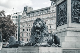 A large bronze lion statue sits majestically on a stone pedestal in an urban environment. Behind the lion, there is a historic building with ornate architectural details, including multiple windows and decorative elements. To the left, trees and modern buildings are visible. The entire scene has a historical yet slightly modern atmosphere.