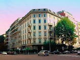 A large, classical-style apartment building with multiple stories, featuring prominent balconies and numerous windows. The structure sits on a street corner, flanked by other residential buildings. A large tree stands to the right, providing some greenery in the urban scene. Several parked cars line the streets, and a pale pastel sky serves as the backdrop.