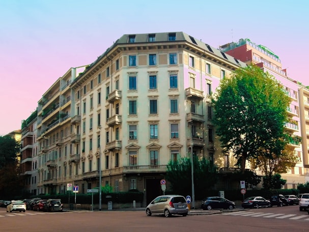A large, classical-style apartment building with multiple stories, featuring prominent balconies and numerous windows. The structure sits on a street corner, flanked by other residential buildings. A large tree stands to the right, providing some greenery in the urban scene. Several parked cars line the streets, and a pale pastel sky serves as the backdrop.