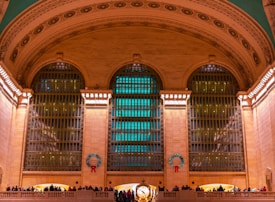 An elegant and grand interior with large arched windows adorned with festive wreaths featuring red ribbons. The stone architecture is highlighted by warm lighting, creating a majestic atmosphere. People are gathered on a balcony, observing the scene or waiting, adding a lively touch.