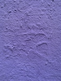 A textured wall painted in a shade of purple. The surface exhibits irregular patterns and roughness characteristic of a plastered finish.