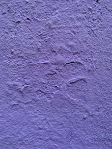 A textured wall painted in a shade of purple. The surface exhibits irregular patterns and roughness characteristic of a plastered finish.