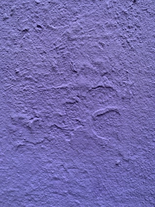 A textured wall painted in a shade of purple. The surface exhibits irregular patterns and roughness characteristic of a plastered finish.
