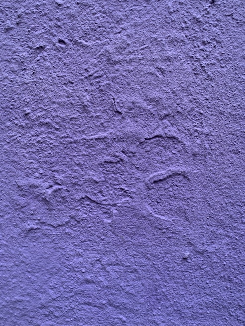 A textured wall painted in a shade of purple. The surface exhibits irregular patterns and roughness characteristic of a plastered finish.