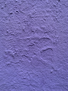 A textured wall painted in a shade of purple. The surface exhibits irregular patterns and roughness characteristic of a plastered finish.