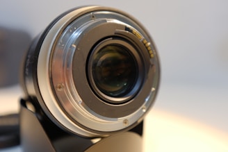A close-up view of a camera lens mount, displaying the metallic and glass components. Screws and connectors are visible around the mount, emphasizing the precision and engineering involved. The background is softly blurred, giving focus to the intricate details of the lens.