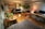 A well-lit studio apartment with wooden flooring, featuring a kitchen area with cabinets, a refrigerator, and a microwave on the left side. A black leather sofa is positioned in the center, adorned with a modern floor lamp beside it. Several potted plants are placed on tables and hanging, offering a green touch to the room. A large desk area has a computer setup, complete with a monitor and laptop, illuminated by a small lamp with a red shade. The ceiling has exposed ductwork, contributing to an industrial aesthetic.