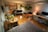 A well-lit studio apartment with wooden flooring, featuring a kitchen area with cabinets, a refrigerator, and a microwave on the left side. A black leather sofa is positioned in the center, adorned with a modern floor lamp beside it. Several potted plants are placed on tables and hanging, offering a green touch to the room. A large desk area has a computer setup, complete with a monitor and laptop, illuminated by a small lamp with a red shade. The ceiling has exposed ductwork, contributing to an industrial aesthetic.