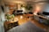 A well-lit studio apartment with wooden flooring, featuring a kitchen area with cabinets, a refrigerator, and a microwave on the left side. A black leather sofa is positioned in the center, adorned with a modern floor lamp beside it. Several potted plants are placed on tables and hanging, offering a green touch to the room. A large desk area has a computer setup, complete with a monitor and laptop, illuminated by a small lamp with a red shade. The ceiling has exposed ductwork, contributing to an industrial aesthetic.
