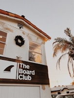 A building with a white facade and a terracotta tile roof features a wooden sign reading 'The Board Club, Newport'. A wreath decorates the upper wall, and a palm tree is visible nearby. The sky is clear with a warm tone, suggesting a sunset or sunrise.