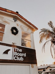 A building with a white facade and a terracotta tile roof features a wooden sign reading 'The Board Club, Newport'. A wreath decorates the upper wall, and a palm tree is visible nearby. The sky is clear with a warm tone, suggesting a sunset or sunrise.