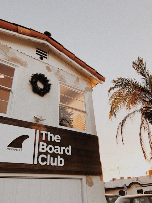 A building with a white facade and a terracotta tile roof features a wooden sign reading 'The Board Club, Newport'. A wreath decorates the upper wall, and a palm tree is visible nearby. The sky is clear with a warm tone, suggesting a sunset or sunrise.